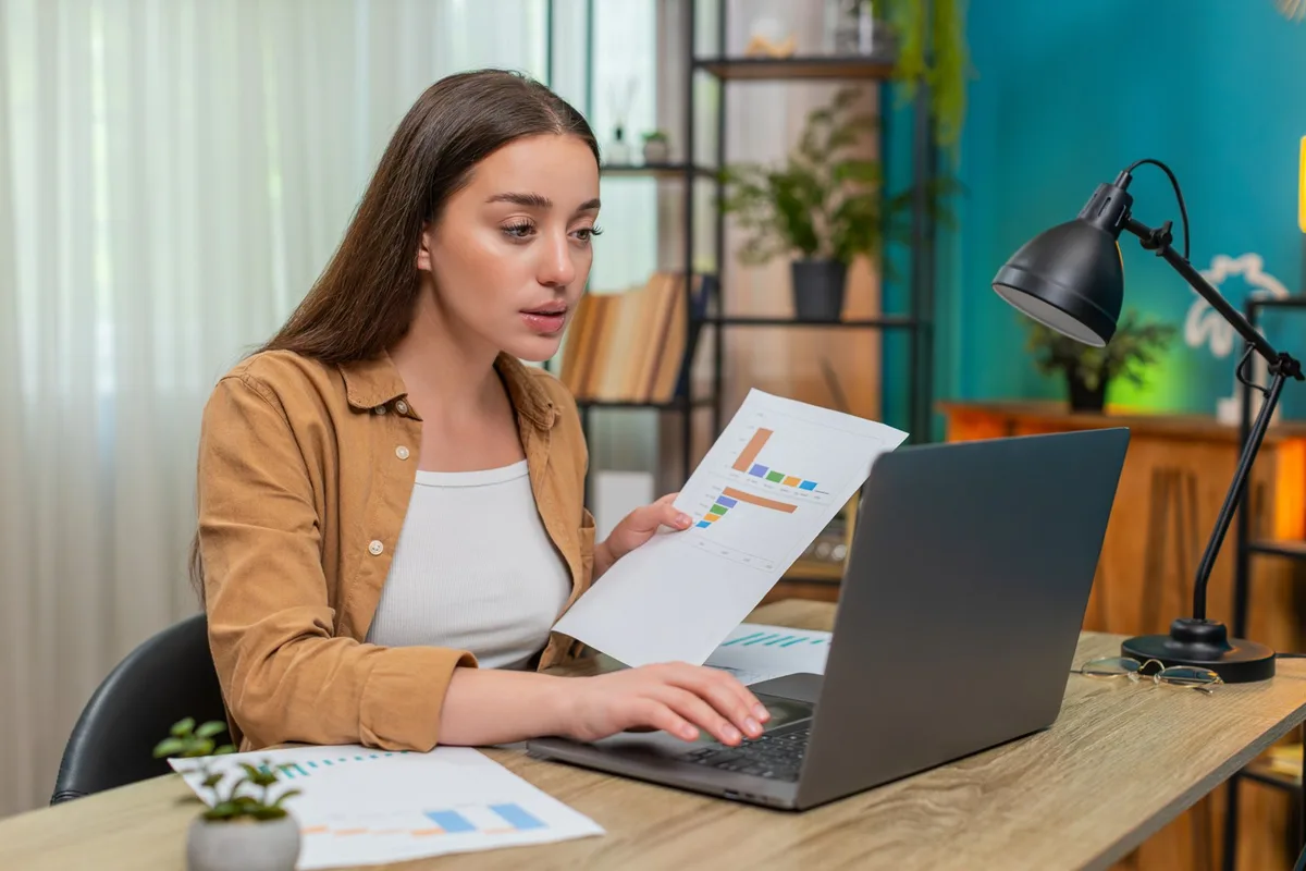 Marketer checking a printed report against the figures on her laptop before it goes to a client